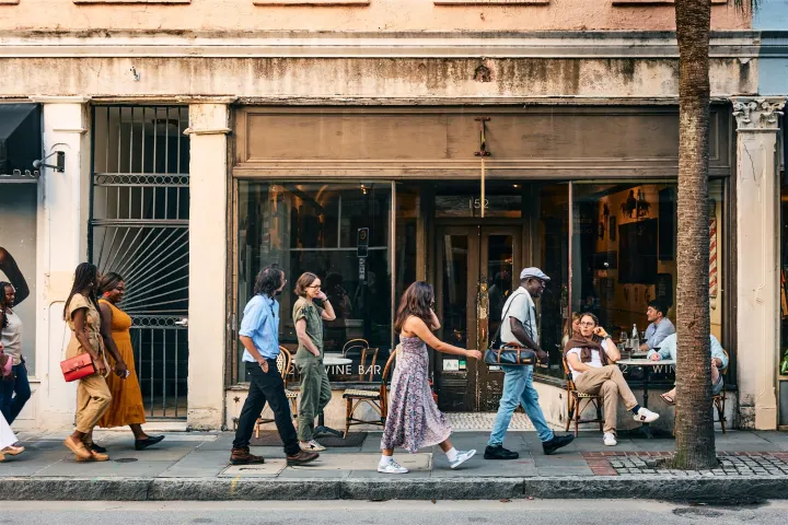 People walk past a vintage storefront with outdoor seating on a sunny day.