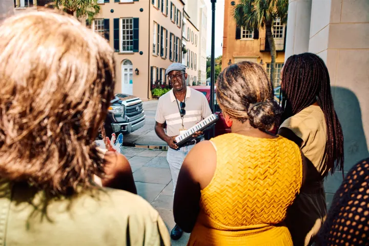 Man playing melodica for a group of people on a sunny street with historic buildings.