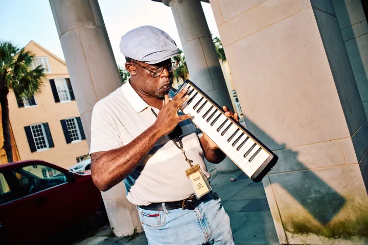 Man playing a melodica outside, wearing a hat and glasses against a building backdrop.