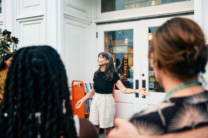 Woman gesturing while speaking to a group outside a bookstore.