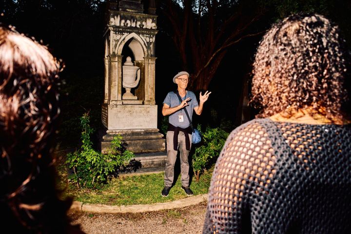Person in hat talks to group near a stone monument at night.