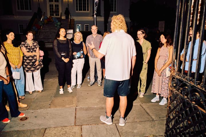 A group of people standing outside a building at night, listening to a man in a white shirt.